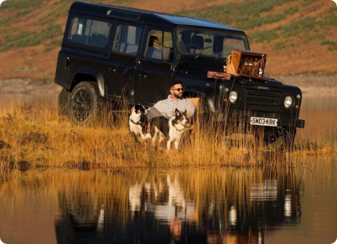 A man sits on the grassy shore of a lake with two dogs beside him, both looking ahead. Behind them is a black off-road vehicle with a wooden Melvill & Moon picnic box displayed on the hood. The lake reflects the scene, creating a mirrored image of the man, dogs, and vehicle. The landscape in the background is a mix of hills and earthy tones.