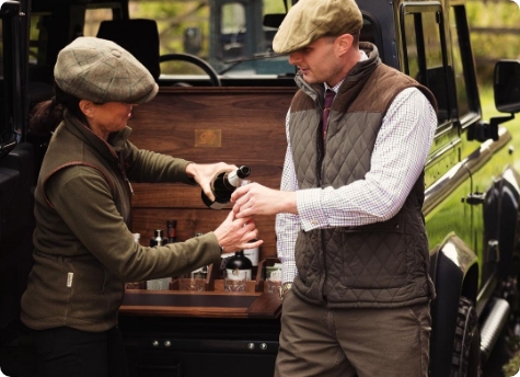 A woman and a man, both wearing traditional outdoor attire with flat caps, stand beside an open vehicle Melvill & Moon bar setup at the back of a black off-road vehicle. The woman is pouring a drink from a bottle, while the man holds out a glass. The setting appears to be outdoors with a grassy background.