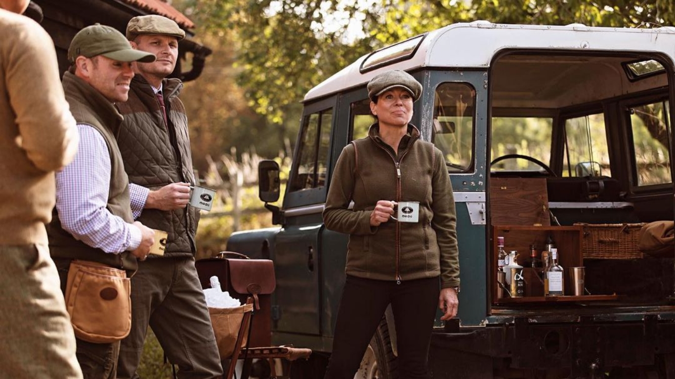 Steffi from Batavia House and a group of people dressed in outdoor attire stand around a classic off-road vehicle with a built-in bar at the rear, displaying Melvill & Moon bottles and cups. The scene appears relaxed. The setting is outdoors with greenery and a rustic building in the background, evoking a countryside gathering.