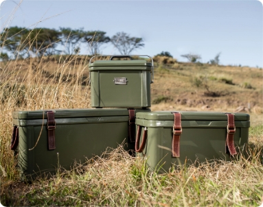 Three Rogue Ice Coolers with leather straps are arranged on dry grassy terrain in a natural setting. The cases are of varying sizes, with one smaller case placed on top of two larger cases. The background shows an open landscape with scattered trees and a clear blue sky.