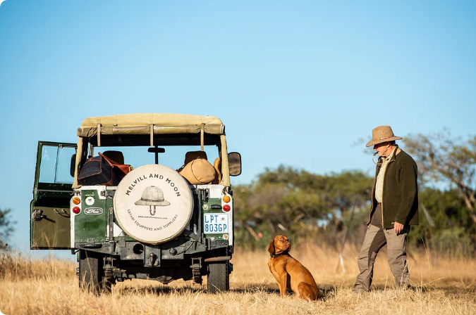 A man wearing a wide-brimmed hat and outdoor attire stands beside a classic off-road vehicle with a "Melvill and Moon" spare tire cover. The vehicle is parked on dry, grassy terrain, and a brown dog sits attentively next to the man. The background features trees and a clear blue sky.